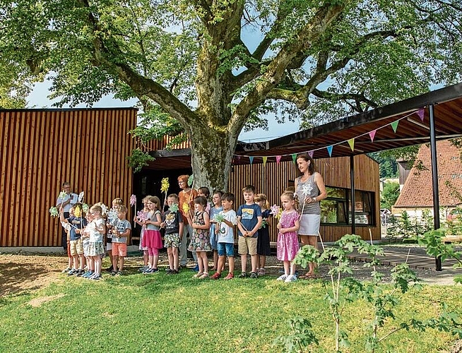 Ein Dankeschön der Kindergärtler: Zusammen mit den Lehrpersonen Käthi Setz (links vom Baum) und Claudia Cecchin profitierten sie vom Schatten des Lindenbaums. Foto: Peter Winkelmann