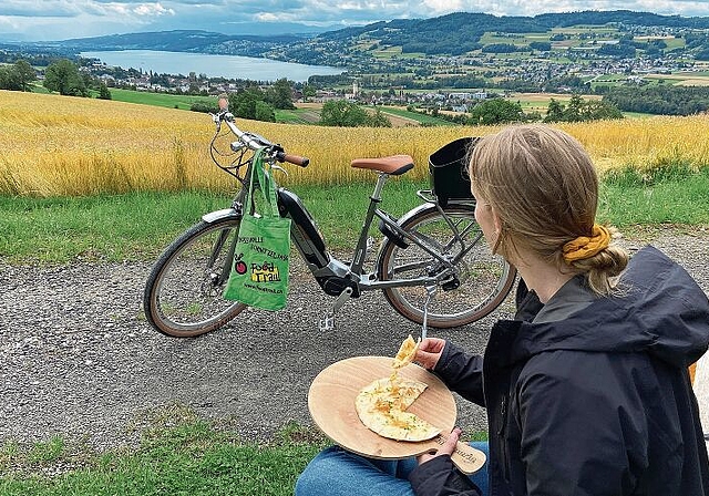 Regionale Köstlichkeiten an den Etappenzielen: FoodTrial-Seetal-E-Bikerin geniesst die Aussicht über den Hallwilersee und das gereichte Häppchen. Foto: zvg
