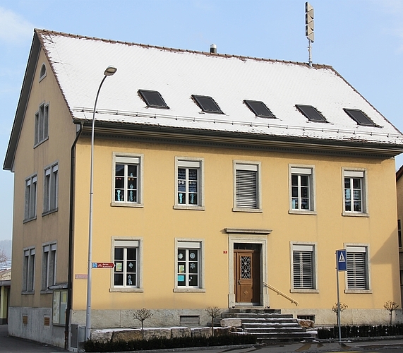 Das 1868 als Geschäftshaus errichtete Gebäude diente von 1875 bis 1958 als Schulhaus: Der spätklassizistische Mauerbau ist in zeittypischer Weise schlicht gestaltet und wirkt durch seine guten Proportionen. Als direktes Gegenüber des Gasthofs Bären und des Bauernhauses Wildegger-strasse 2 kommt dem Gebäude erheblicher Situationswert zu.
