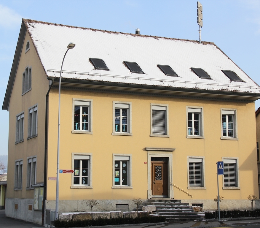 Das 1868 als Geschäftshaus errichtete Gebäude diente von 1875 bis 1958 als Schulhaus: Der spätklassizistische Mauerbau ist in zeittypischer Weise schlicht gestaltet und wirkt durch seine guten Proportionen. Als direktes Gegenüber des Gasthofs Bären und des Bauernhauses Wildegger-strasse 2 kommt dem Gebäude erheblicher Situationswert zu.

